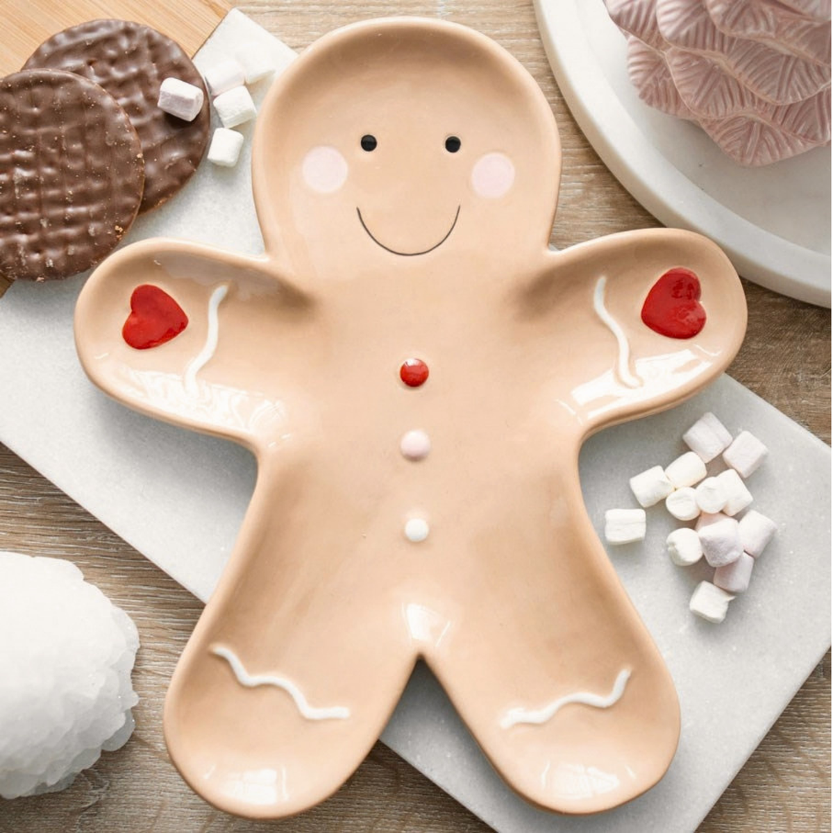 A gingerbread man shaped ceramic serving plate with a smiling face, red heart buttons, and white icing details, styled on a wooden table with cookies and marshmallows.