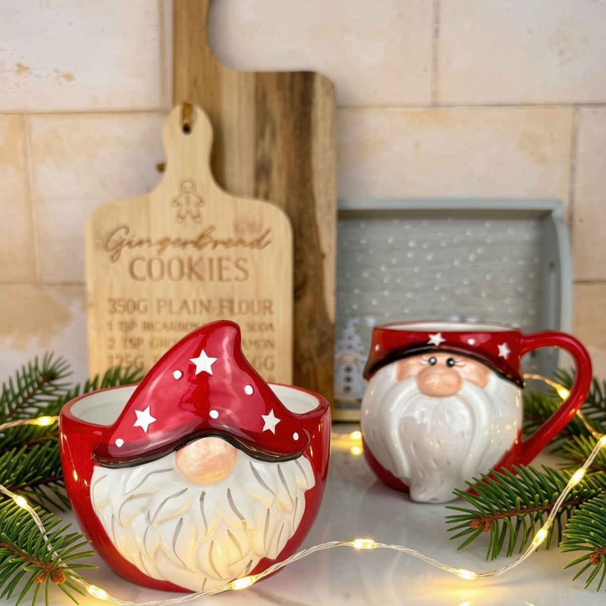 Christmas gonk themed ceramic bowl styled on a countertop with pine branches and warm fairy lights, with a wooden cutting board and tiled backsplash in the background.