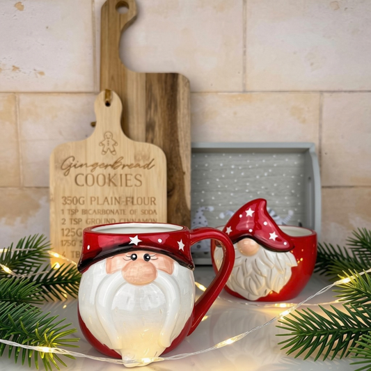 Red and white ceramic Christmas gonk mug styled on a countertop with pine branches, fairy lights, a wooden cutting board, and a tiled wall background.