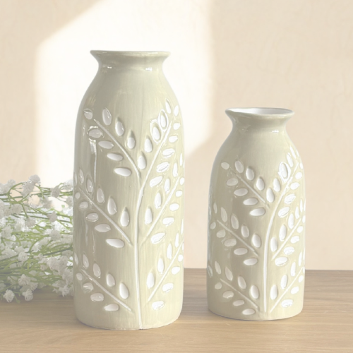 Two ceramic vases with a leafy branch pattern, one tall and one short, in a light beige color, displayed on a wooden surface against a warm beige background.