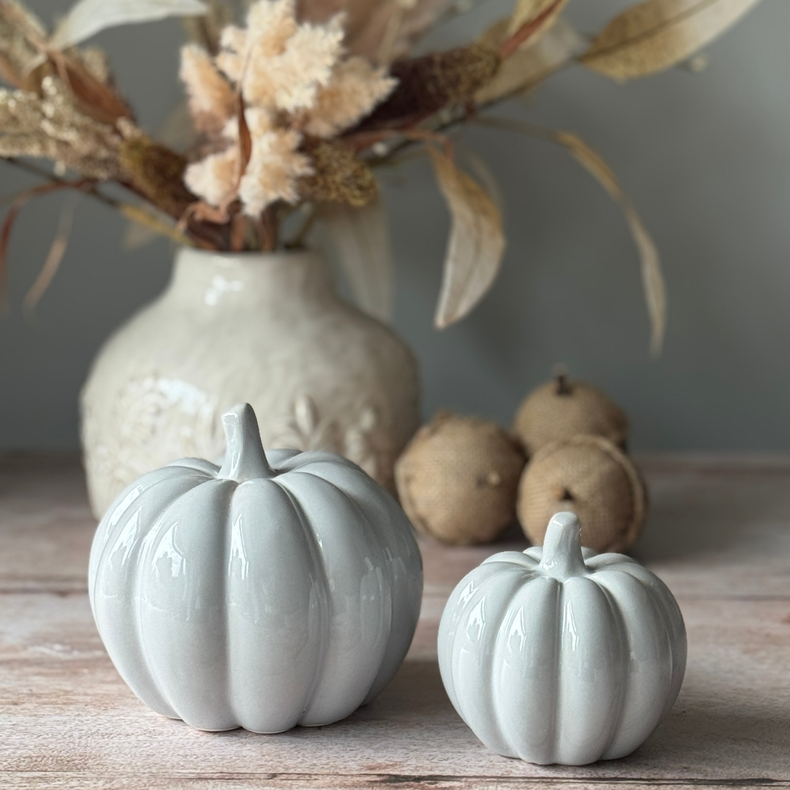 Two grey ceramic pumpkins on a wooden surface with a vase and acorns in the background.