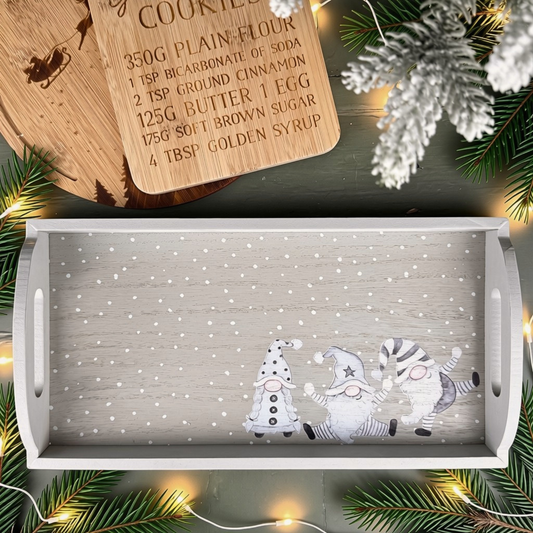A rectangular wooden serving tray with a snowy white-grey grained surface and a printed illustration of three gnomes at the bottom, styled on a dark tabletop with pine branches, fairy lights, a wooden recipe card, and white snow-dusted evergreen accents.