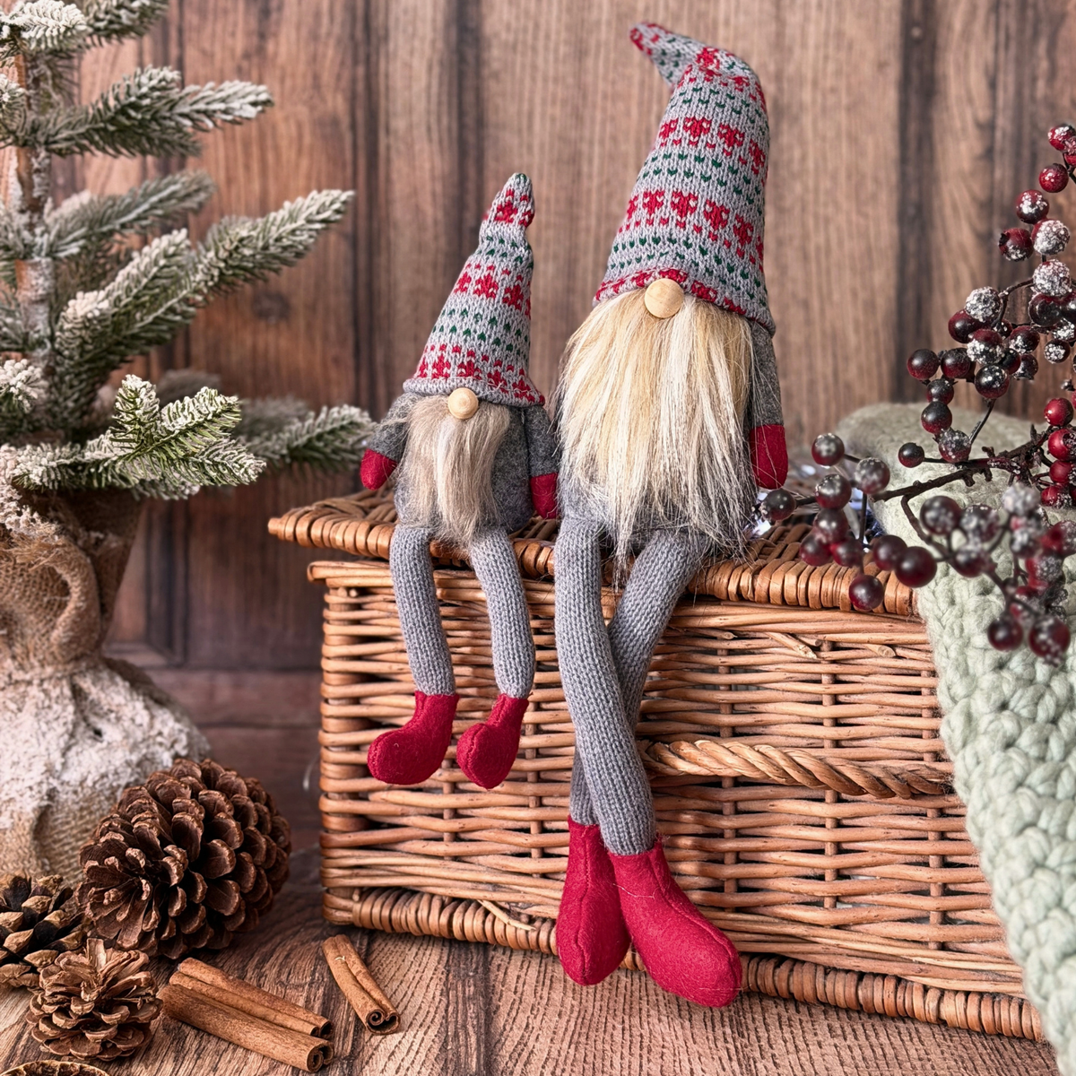 Two knitted Christmas gonkswith long blonde yarn hair, grey knit bodies, patterned grey hats with red motifs, and red felt boots, posed on a wicker basket with rustic holiday decor in the background.