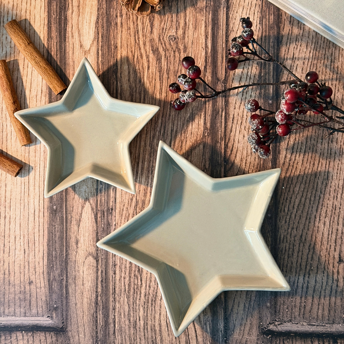 Two star-shaped ceramic dishes in a light grey glaze, arranged on a rustic wooden tabletop with cinnamon sticks and red berries as decorative props.