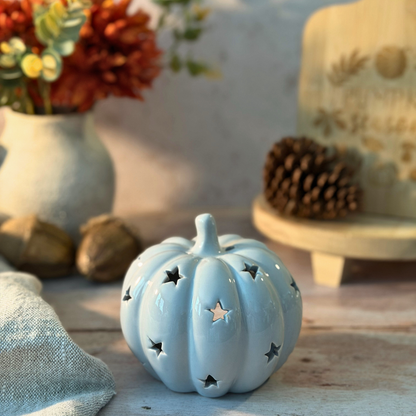 A small glossy light grey decorative pumpkin with star-shaped cutouts, styled on a rustic tabletop with autumn props including a vase of flowers, pinecone, acorns, and a fabric cloth.