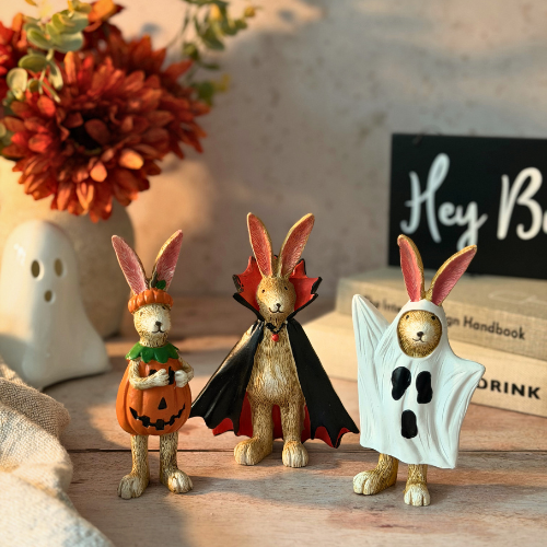 Three small Halloween rabbit figurines standing on a wooden surface: one in an orange pumpkin outfit, one as a vampire with a black cape, and one in a white ghost costume. Decorative flowers, a ghost-shaped object, and books are blurred in the background.