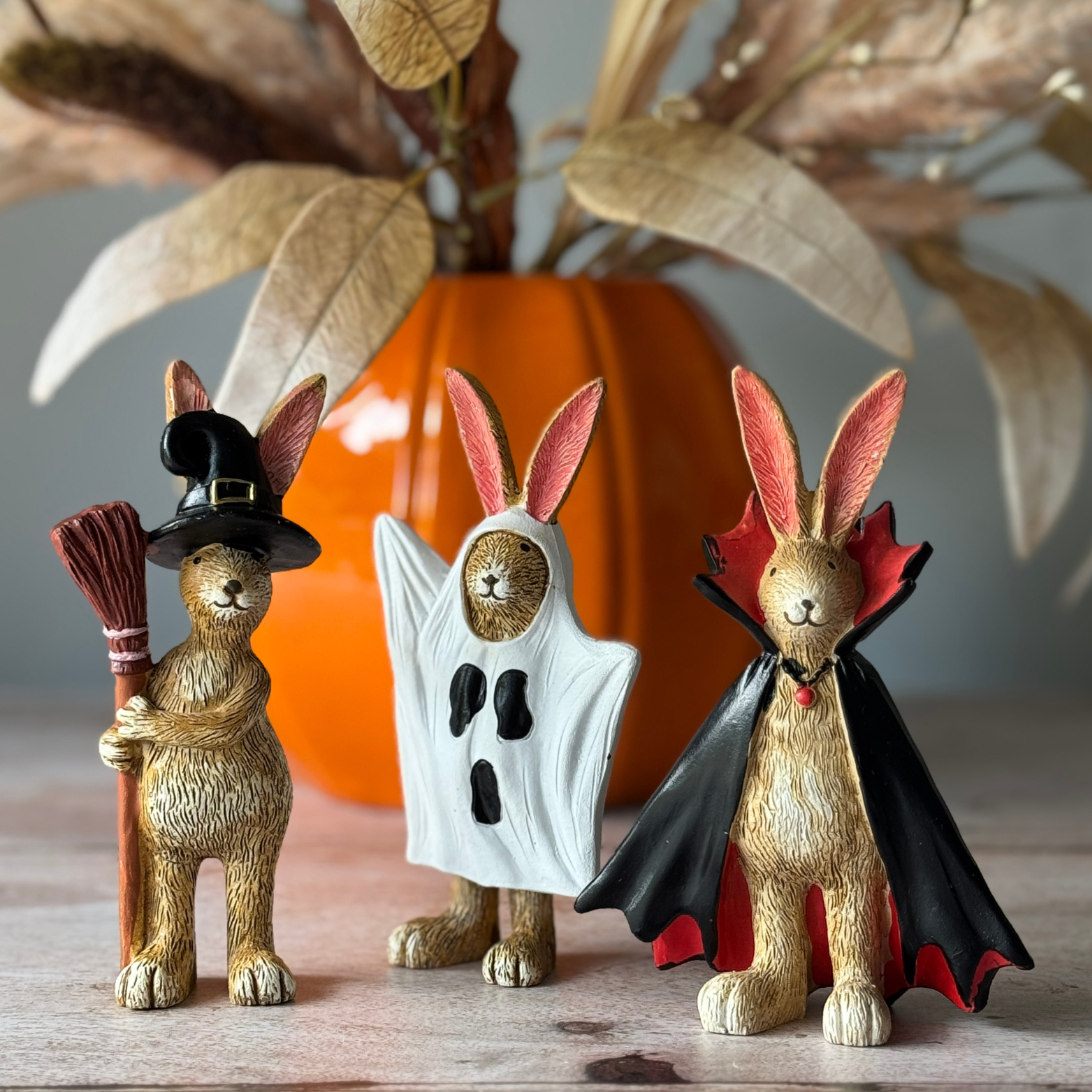 Three rabbit figurines dressed as a witch, ghost, and vampire with a pumpkin in the background.