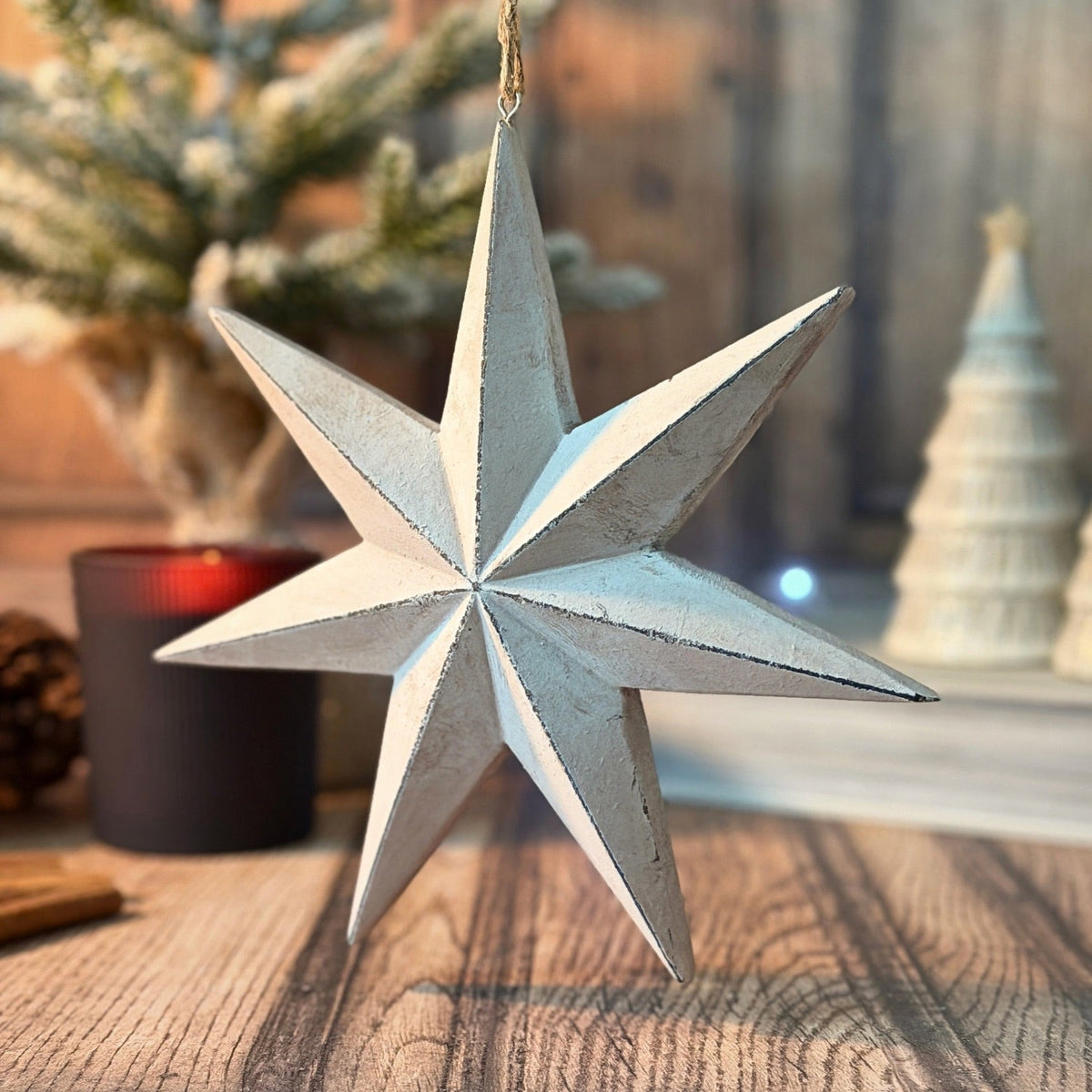 A rustic six-pointed star Christmas ornament with a distressed white and light blue finish, hanging on a wooden table with a blurred holiday background.