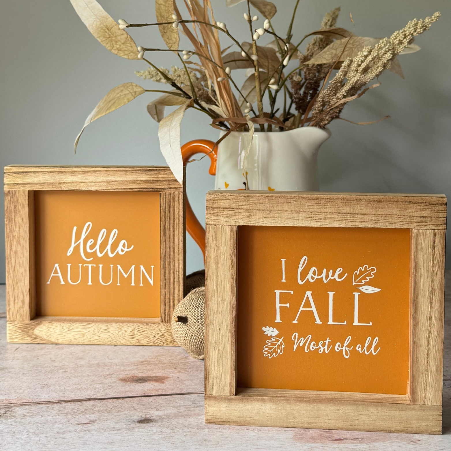 Two wooden picture frames with autumn-themed text on an orange background, placed in front of a vase with dried plants.