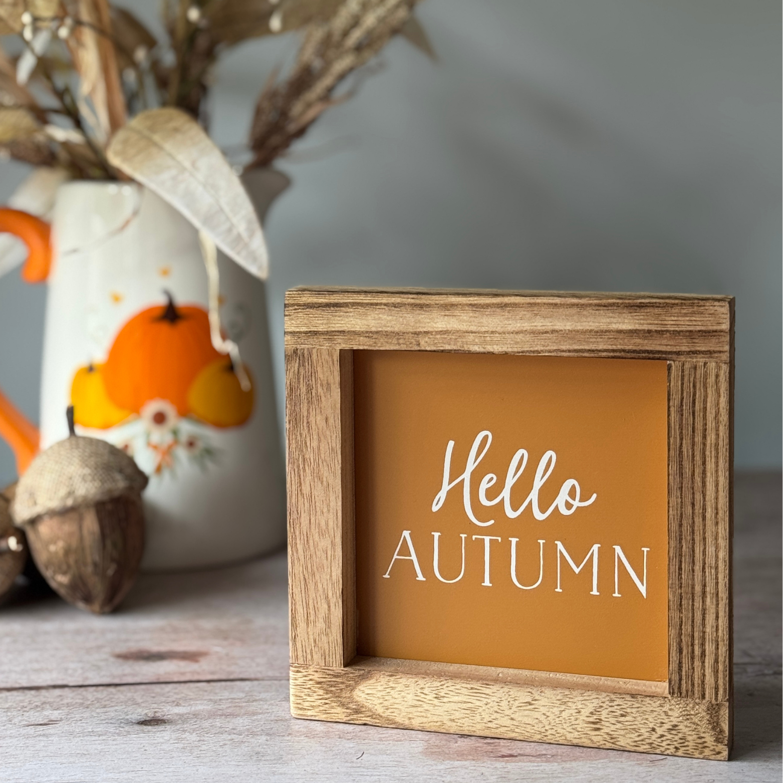 Wooden sign with 'Hello Autumn' text in front of a decorative vase with pumpkins and acorns.