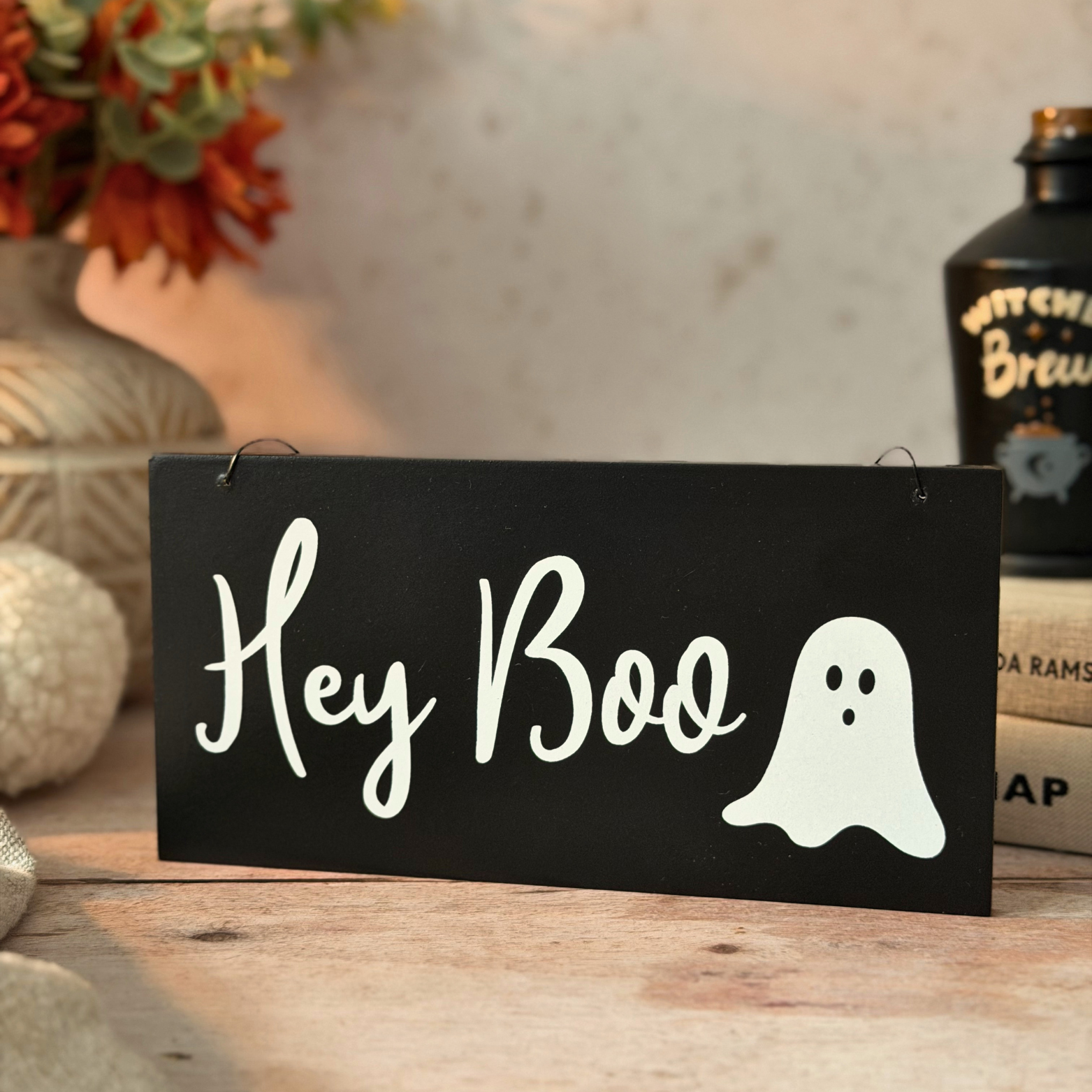 A rectangular black sign with white script reading 'Hey Boo' and a white ghost graphic, displayed on a wooden surface in a styled indoor setting with autumn decor and books in the background.