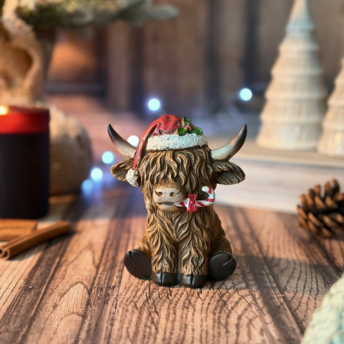 A Highland cow figurine decorated with a red Santa hat and a red-and-white candy cane, sitting on a wooden surface with a warm, festive background.