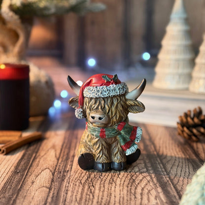 A small Highland cow figurine wearing a red Santa hat and a green and red striped scarf, sitting on a wooden surface with a warm, festive background.