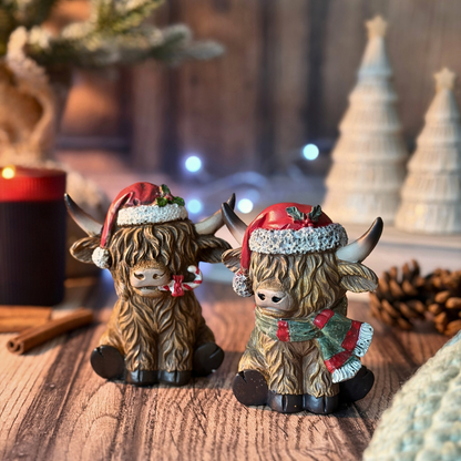 Two Highland cow figurines decorated for Christmas, sitting on a wooden surface with a blurred festive background containing candles, pinecones, and holiday lights.