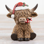 Festive figurine of a Highland Cow wearing a Santa hat with a candy cane in his mouth on a wooden surface