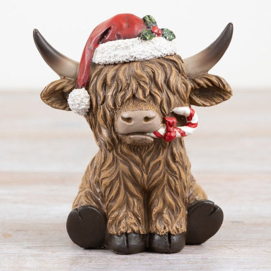 Festive figurine of a Highland Cow wearing a Santa hat with a candy cane in his mouth on a wooden surface