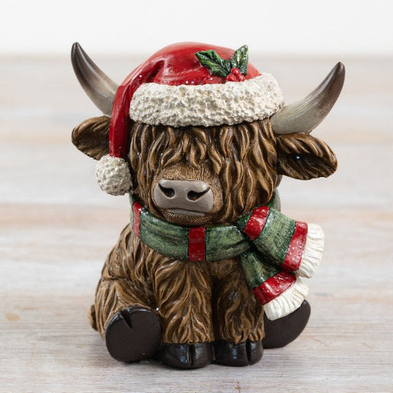 Decorative figurine of a Highland cow wearing a Santa hat and scarf on a wooden surface.