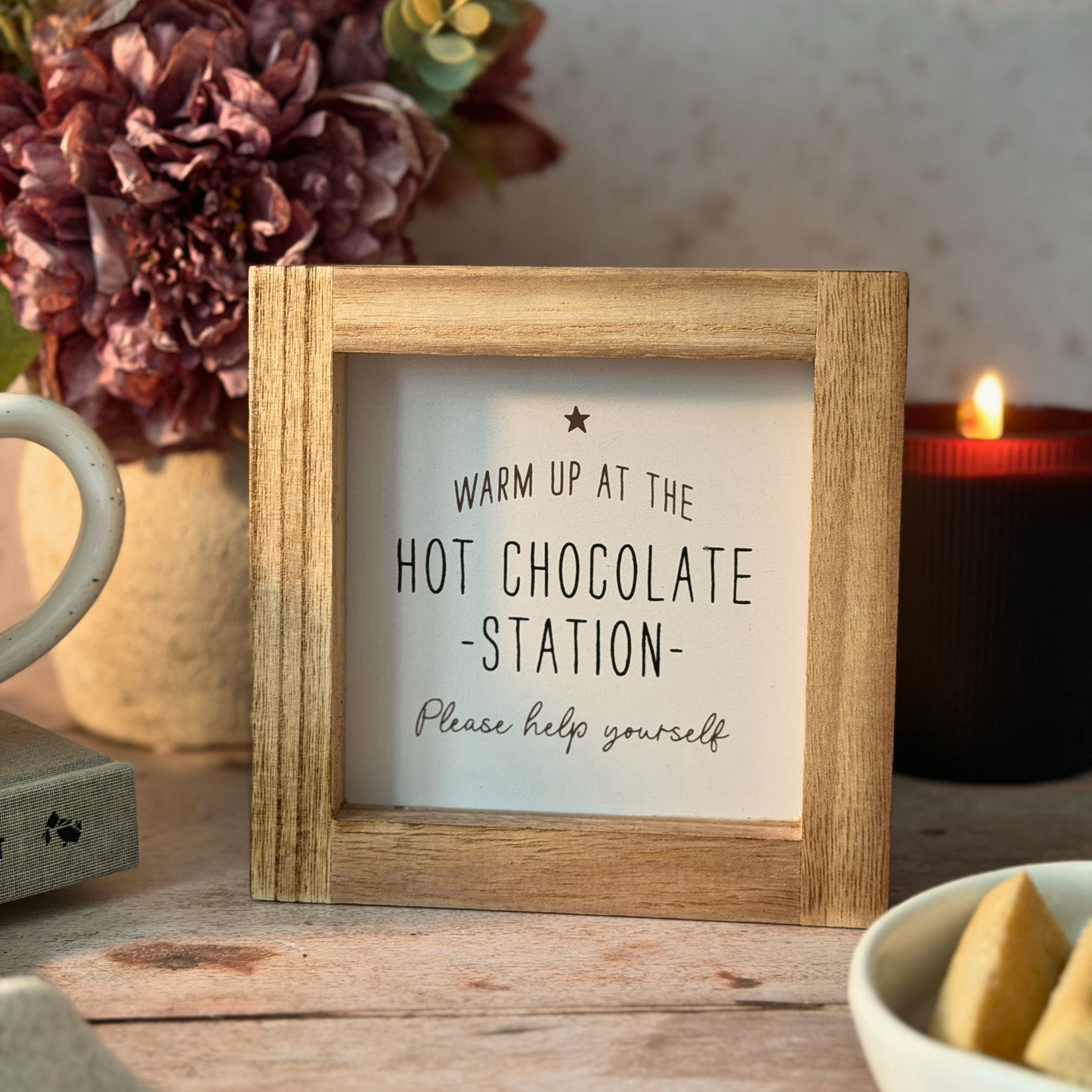 A small square wooden picture frame sign with a humorous quote about hot chocolate, displayed on a rustic wooden table with a mug, flowers, and a candle in the background.