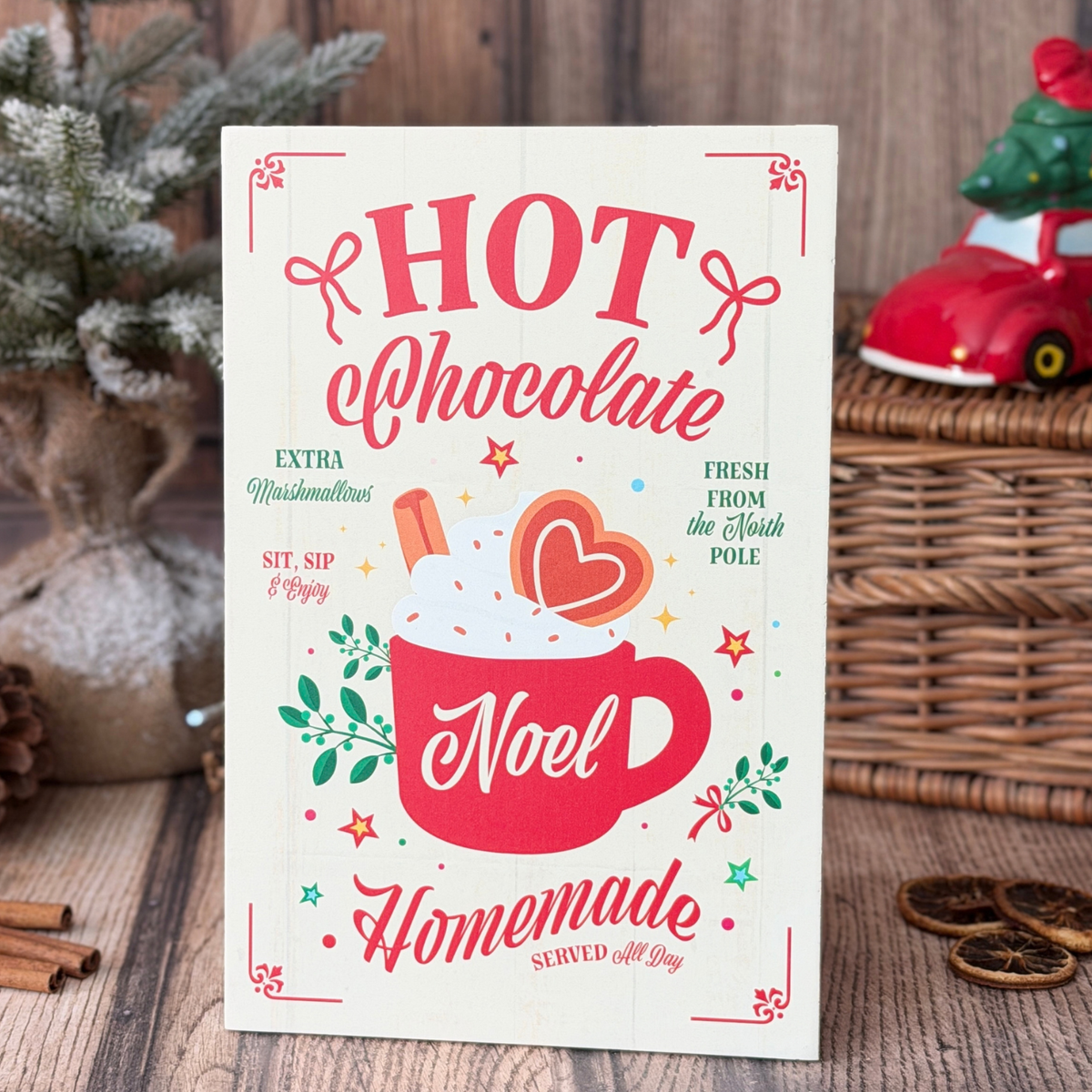A decorative sign featuring a red mug with whipped cream, cinnamon stick, and heart illustration, surrounded by festive text reading 'HOT Chocolate' on a rustic wooden tabletop with holiday props.