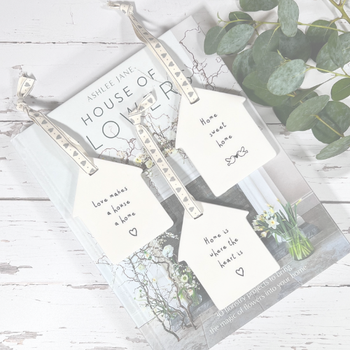 Three house-shaped paper tags with silver string and heart-patterned ribbons are arranged on a book over a white wood surface, with green eucalyptus leaves in the corner. The tags have handwritten phrases including 'love makes a home a home' and 'home is where the heart is'.