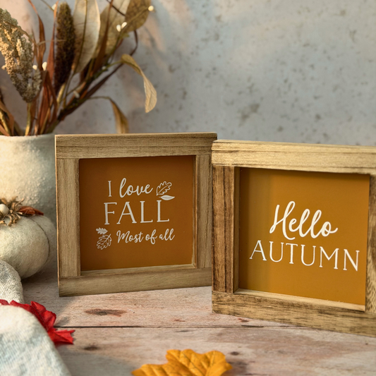 Two small square wooden sign plaques with autumn-themed quotes, displayed on a rustic wooden surface with fall decor props including a pumpkin, dried flowers, and leaves.
