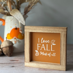 Wooden frame with 'I love Fall Most of all' text on an orange background, placed on a surface with decorative items.