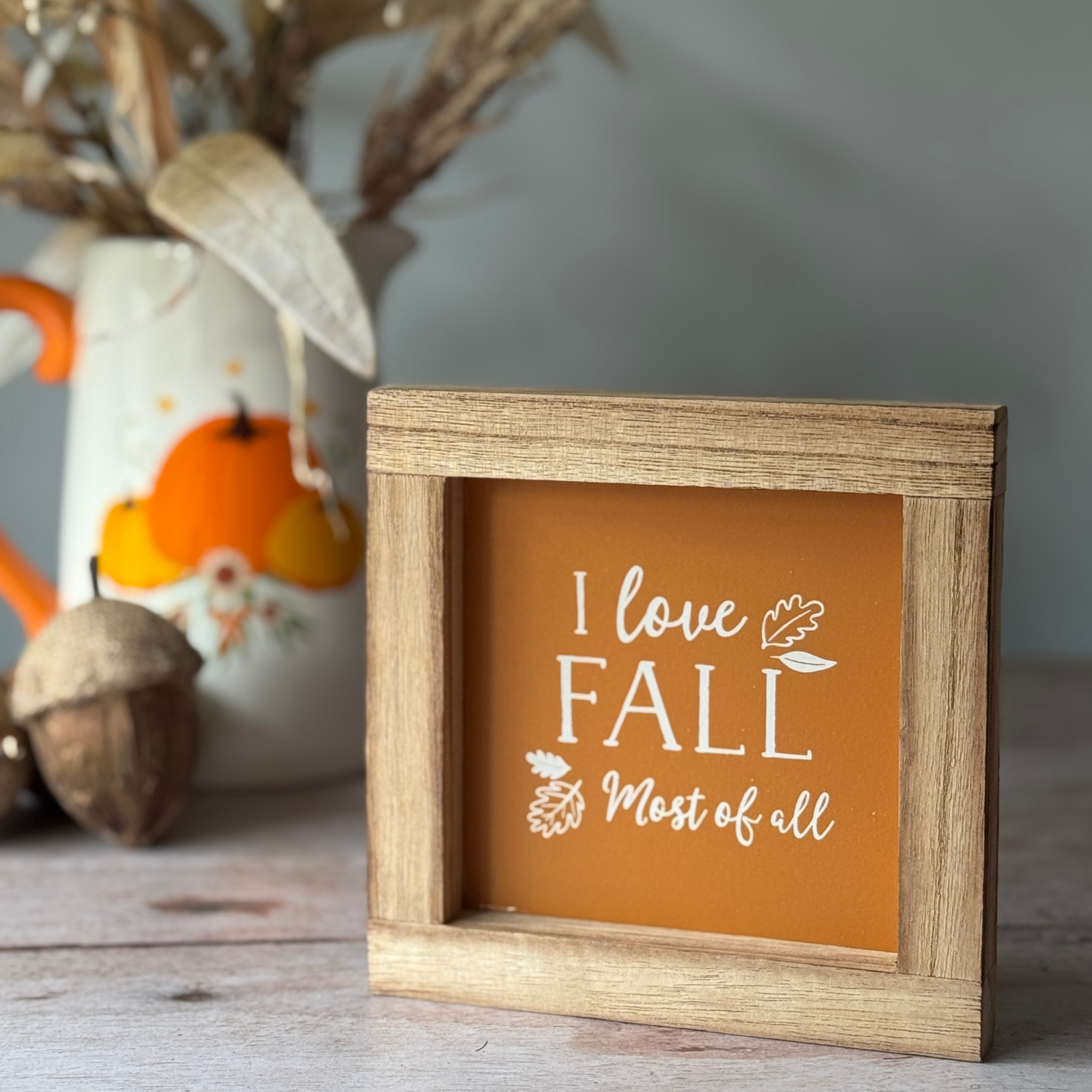 Wooden frame with 'I love Fall Most of all' text on an orange background, placed on a surface with decorative items.