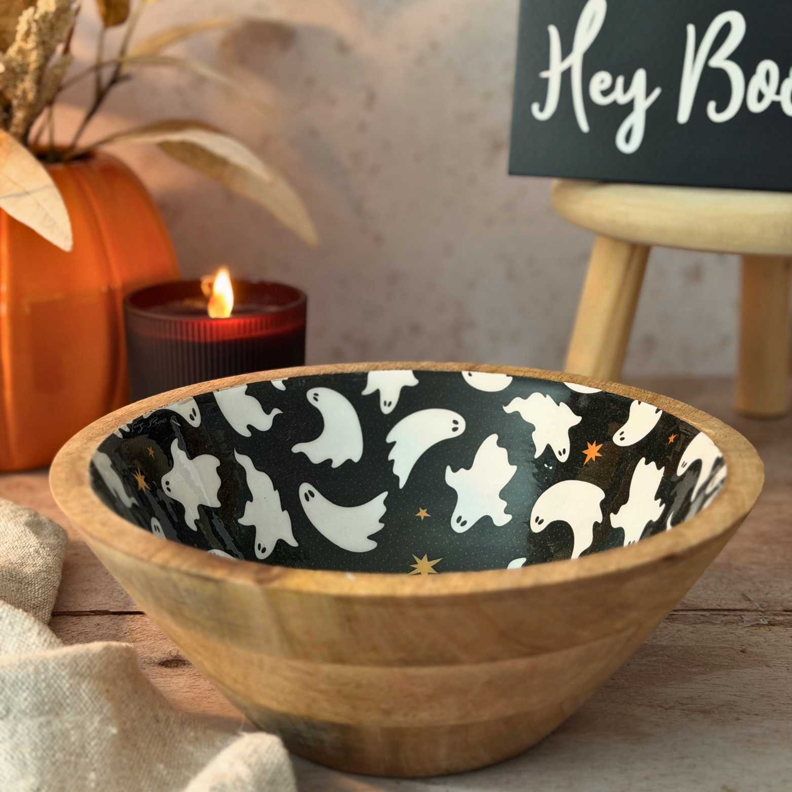 Wooden bowl with a black and white ghost pattern on the inside, styled on a tabletop with a lit candle, an orange pumpkin, and a sign in the background.