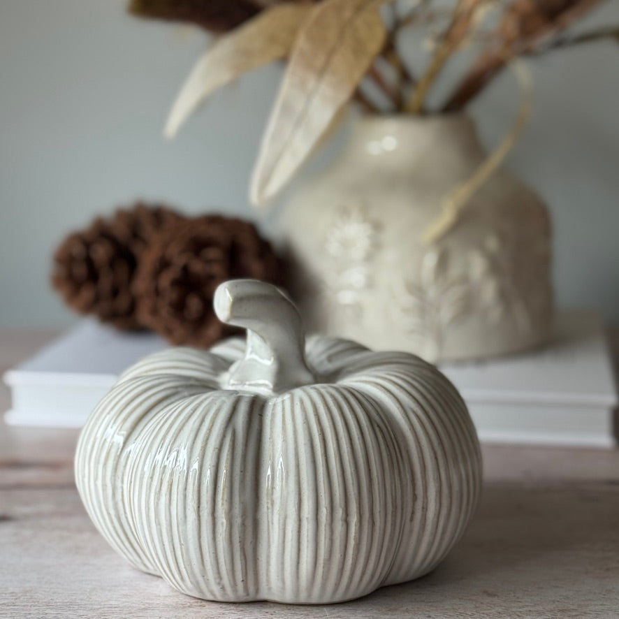 White ceramic pumpkin on a surface with a blurred vase and pinecones in the background