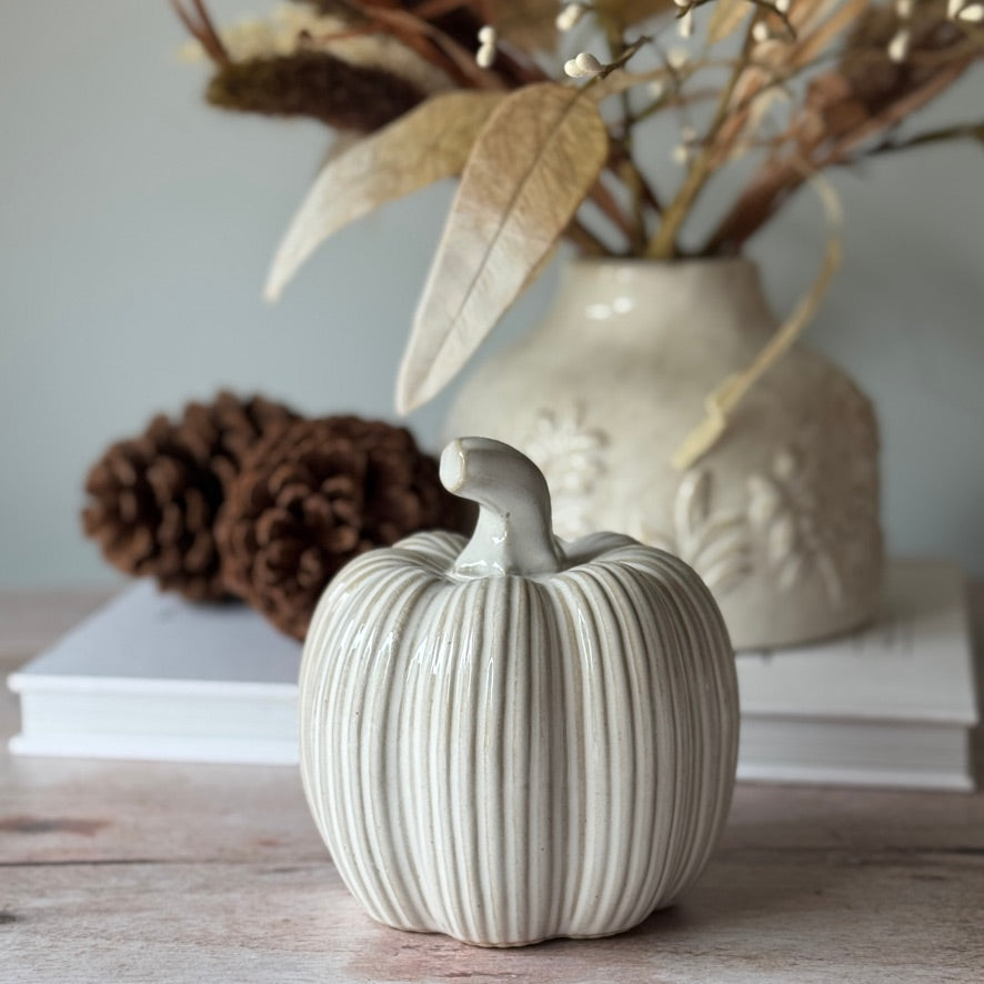 White ceramic pumpkin on a wooden surface with a vase and pinecones in the background.