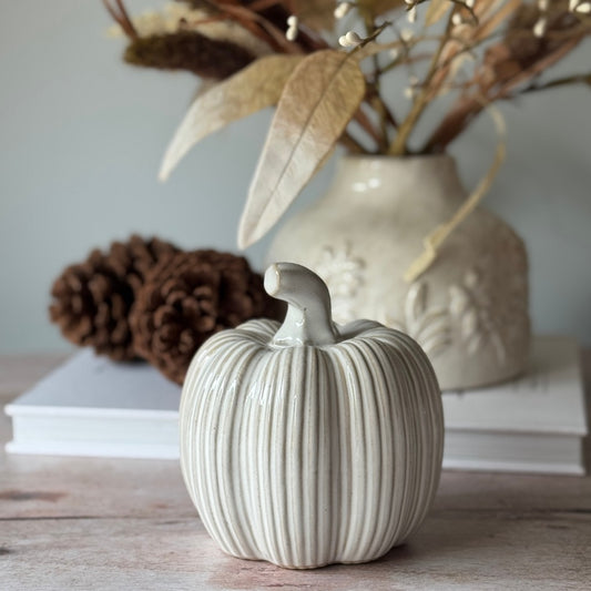 White ceramic pumpkin on a wooden surface with a vase and pinecones in the background.