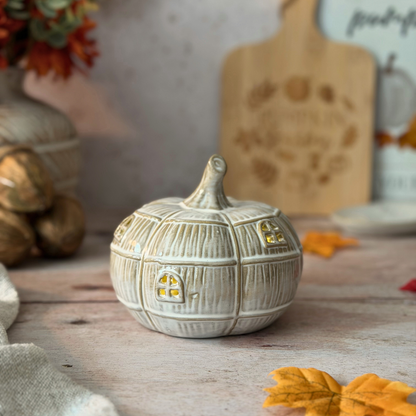 A small decorative ceramic pumpkin figurine with a textured paneled surface, yellow window details, and a short stem, displayed on a rustic wooden surface with autumn leaves and fall-themed decor in the background.