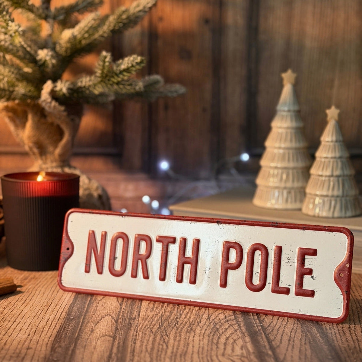 A rectangular vintage-style metal sign reading “NORTH POLE” in raised red lettering on an off-white background, styled on a wooden surface with a lit candle, a small tree, and two Christmas tree ornaments in the background.