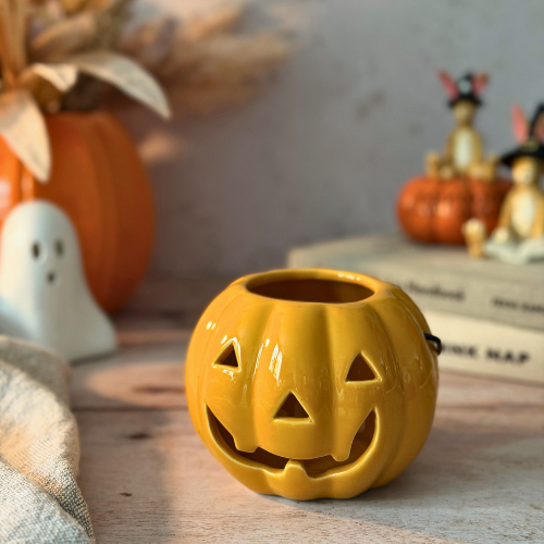 A small orange jack-o'-lantern-shaped candy bucket with a carved face sits on a wooden surface in a styled autumn setting with pumpkins, a ghost figurine, books, and bunny decorations in the background.