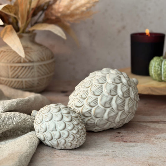 Two decorative faux pinecones in a textured, matte off-white finish, styled on a rustic wooden surface with a linen cloth, a woven vase with dried grass, and a lit black candle in the background.