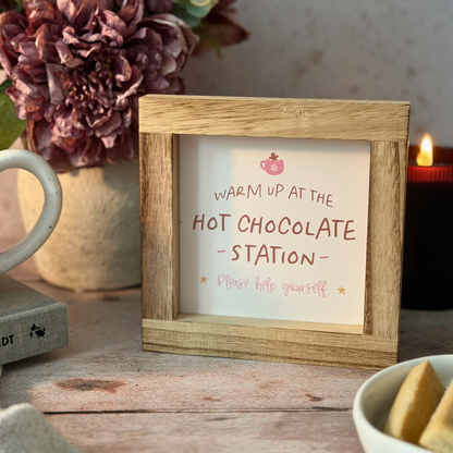 A small wooden picture frame holding a sign that reads 'WARM UP AT THE HOT CHOCOLATE STATION Please help yourself' with a small pink hot chocolate icon, styled on a rustic wooden table with a mug, flowers, and a candle in the background.