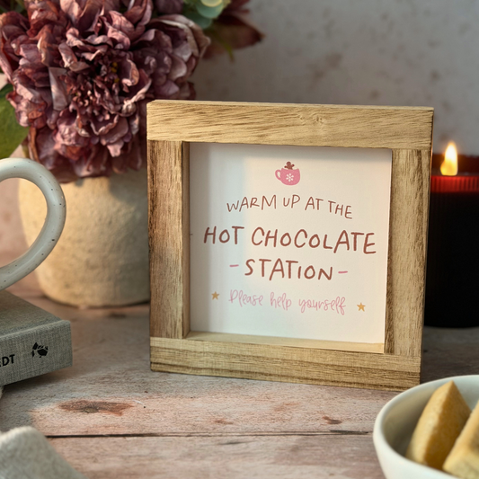 A small wooden picture frame holding a sign that reads 'WARM UP AT THE HOT CHOCOLATE STATION Please help yourself' with a small pink hot chocolate icon, styled on a rustic wooden table with a mug, flowers, and a candle in the background.
