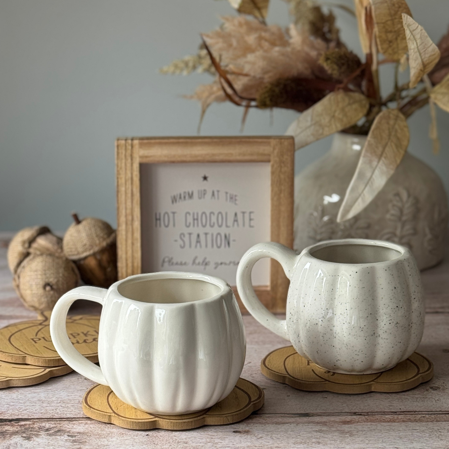 Two ceramic mugs on coasters with a decorative sign and vase in the background.