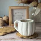 White ceramic pumpkin mug on a wooden coaster, surrounded by autumn-themed decor.
