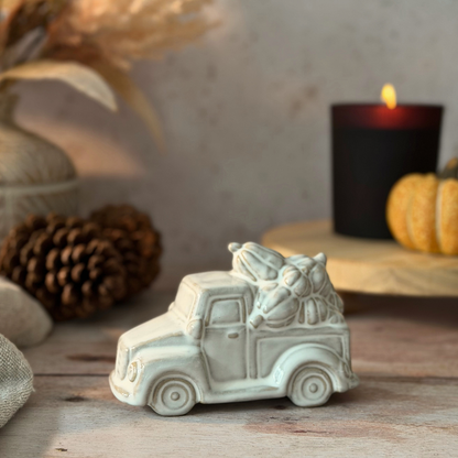A small off-white ceramic Christmas truck figurine carrying wrapped gifts, styled on a rustic wooden surface with a lit black candle, pinecones, and dried grass in the background.