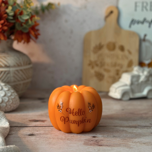 A small orange pumpkin-shaped candle with a lit flame sits on a rustic wooden surface. The front of the candle reads “Hello Pumpkin” in dark script, with small decorative leaf motifs on either side.