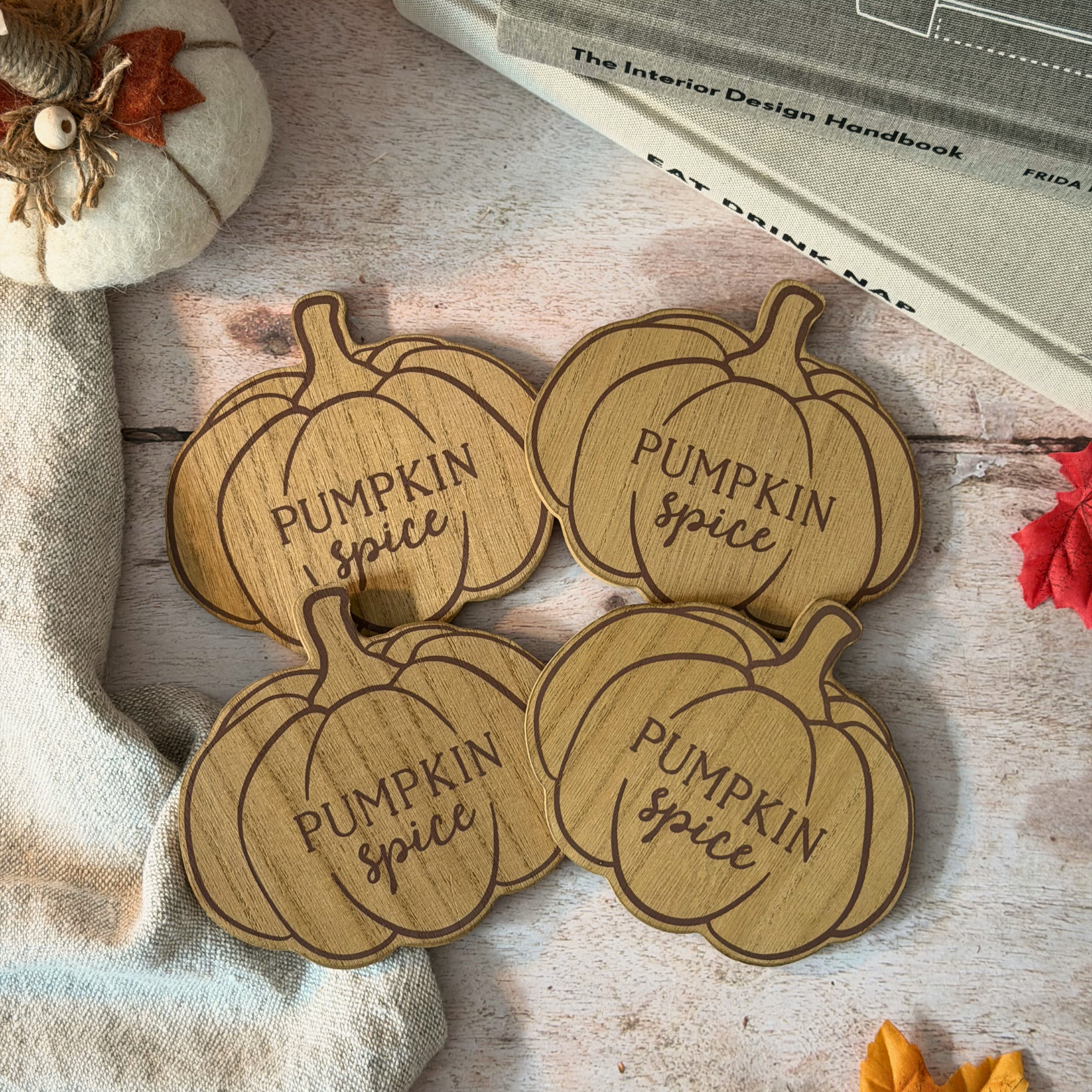 Four pumpkin-shaped wooden coasters engraved with 'Pumpkin Spice' in a warm autumnal setting, surrounded by a small decorative pumpkin, fabric, and books.