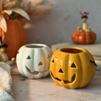 Two small pumpkin-shaped ceramic candle holders with carved jack-o'-lantern faces, one in matte white and one in orange styled on a wooden surface with autumn decor in the background.