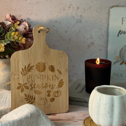 A light natural wood cutting board with a hanging loop, engraved with 'Pumpkin Season' and autumn motifs, styled on a rustic table with a lit candle, a white ceramic pumpkin cup, and dried flowers in the background.