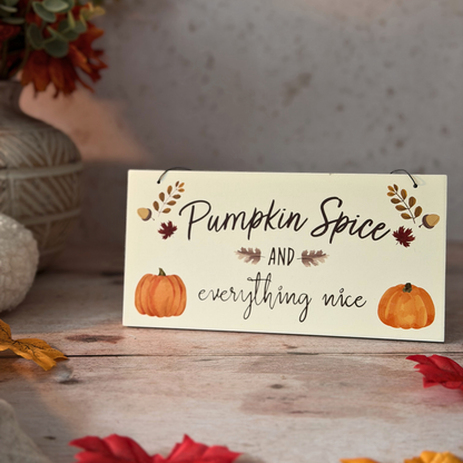 A decorative sign with the phrase “Pumpkin Spice and everything nice” in black script, decorated with small orange pumpkins, leaves, and acorns, displayed on a rustic wooden surface with fall leaves and a vase in the background.