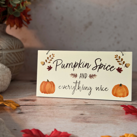A decorative sign with the phrase “Pumpkin Spice and everything nice” in black script, decorated with small orange pumpkins, leaves, and acorns, displayed on a rustic wooden surface with fall leaves and a vase in the background.