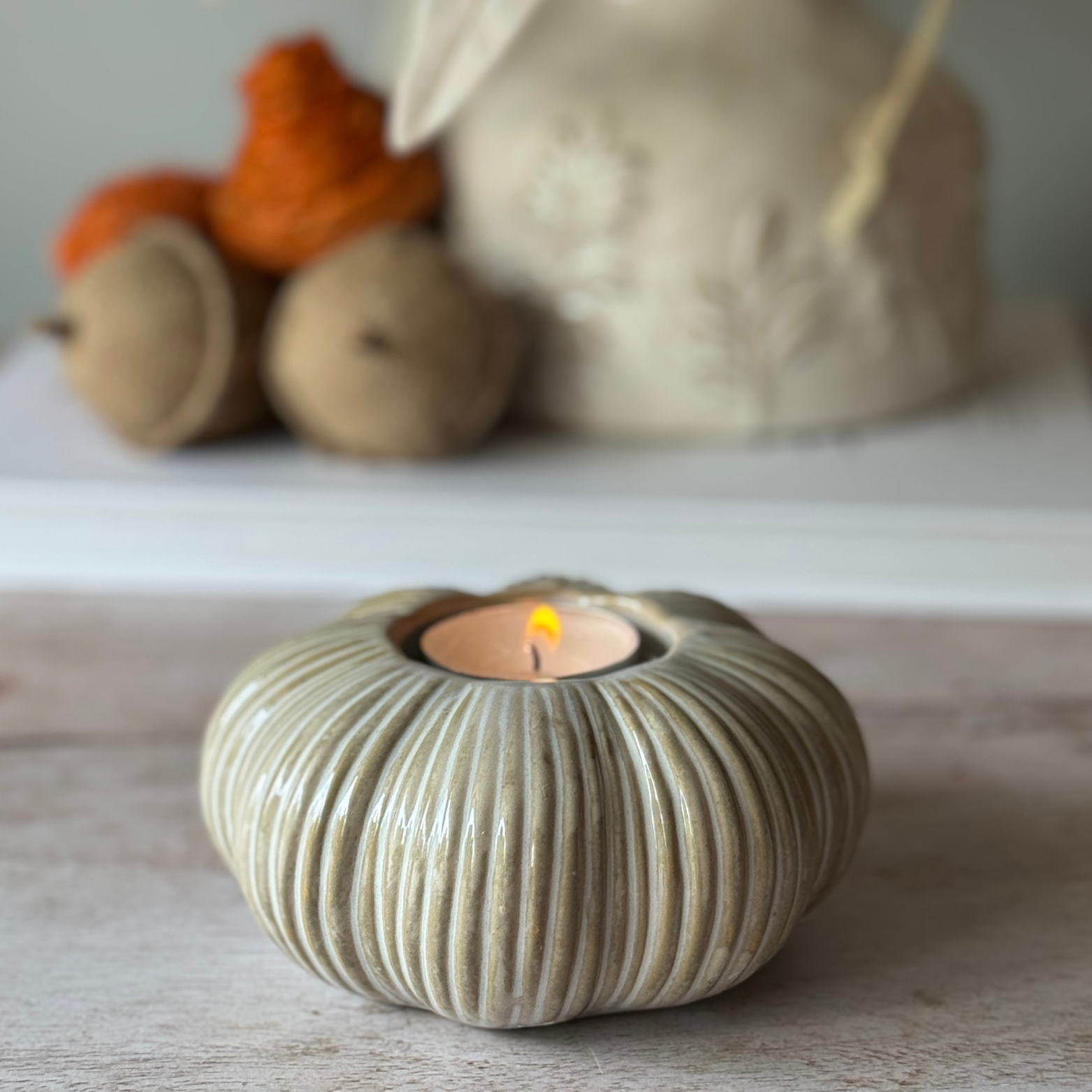 Ceramic tealight holder with a lit candle on a wooden surface, with blurred decorative items in the background.