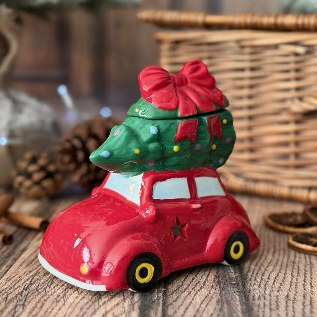 A oil burner styled as a red car carrying a green Christmas tree with a red bow, set on a rustic wooden surface with a wicker basket and pinecones in the background.