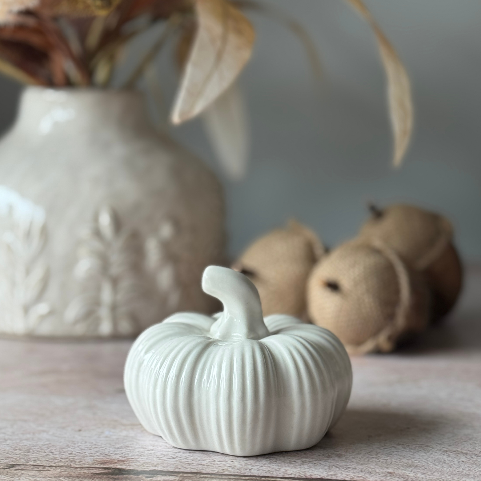White ceramic pumpkin on a wooden surface with a blurred vase and acorns in the background.