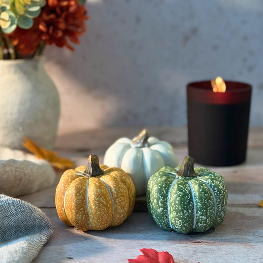 Three decorative miniature pumpkins in yellow, white, and green are arranged on a rustic wooden surface next to a lit black candle and a vase of autumn flowers.